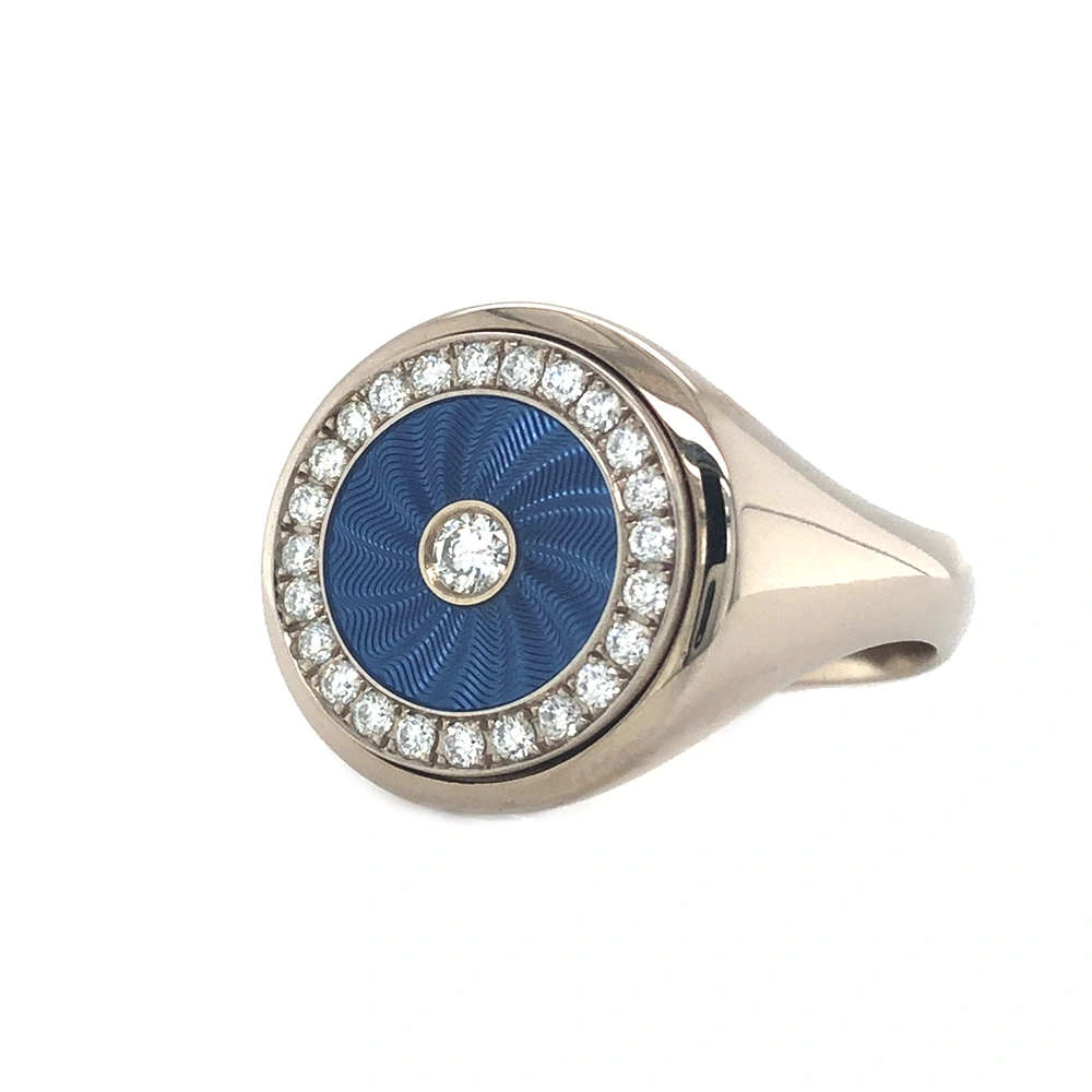 Eleganter runder Ring aus Weißgold mit transluzider hellblauer Emaille und feinem Wellenmuster. Das Inlay ist von 25 funkelnden Brillanten eingefasst. Die Ringschiene ist unterhalb des Ringkopfes tailliert und auf der Unterseite verbreitert, was Komfort und eine markante Form verleiht.